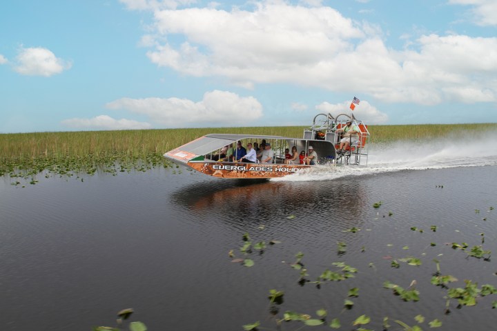 Airboat speeding through a marsh with people on board under a cloudy sky.