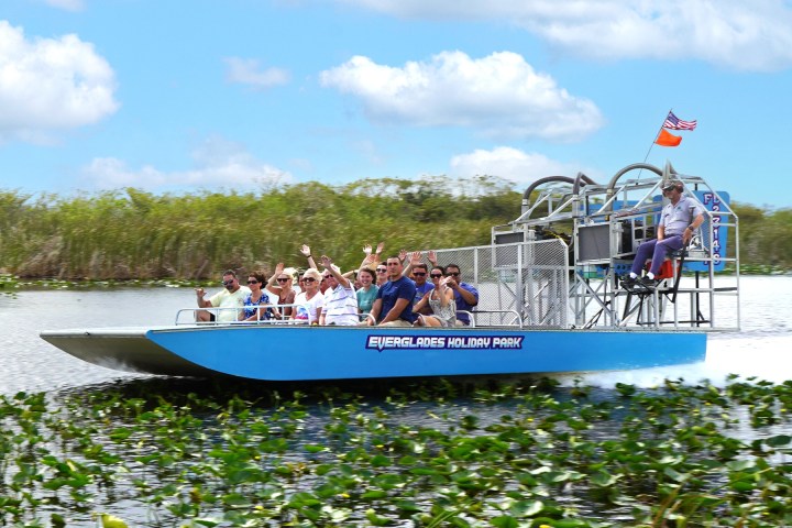 Airboat with tourists speed through wetlands under a blue sky.
