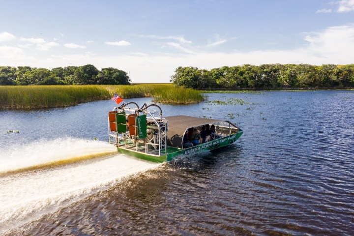 Airboat cruising on a wide lake with grassy banks and trees under a clear blue sky.