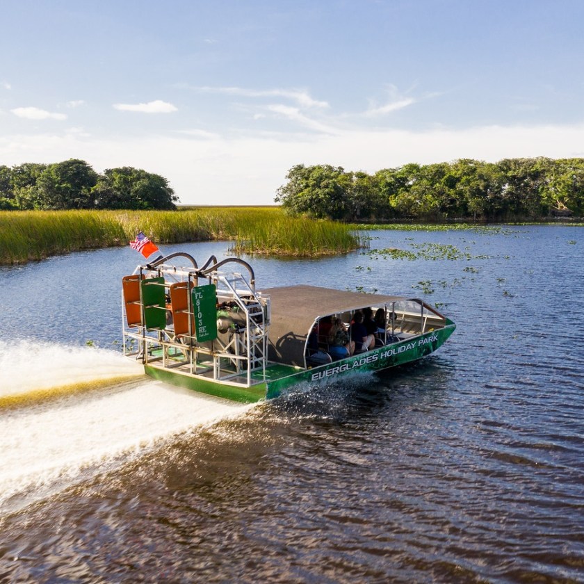 Airboat cruising on a wide lake with grassy banks and trees under a clear blue sky.