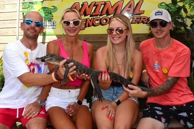 Four smiling people holding a small alligator on a bench with an 'Animal Encounters' sign above.