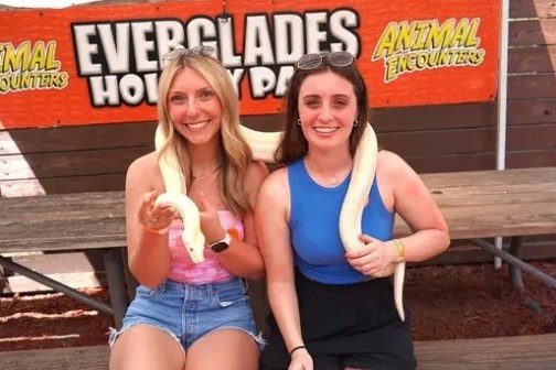 Two women holding snakes with 'Everglades Animal Encounters' sign in background.