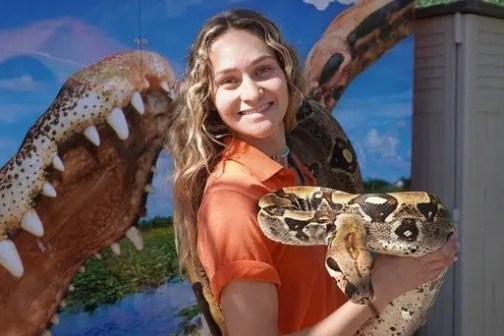 Woman smiling and holding a snake with crocodile image in background.