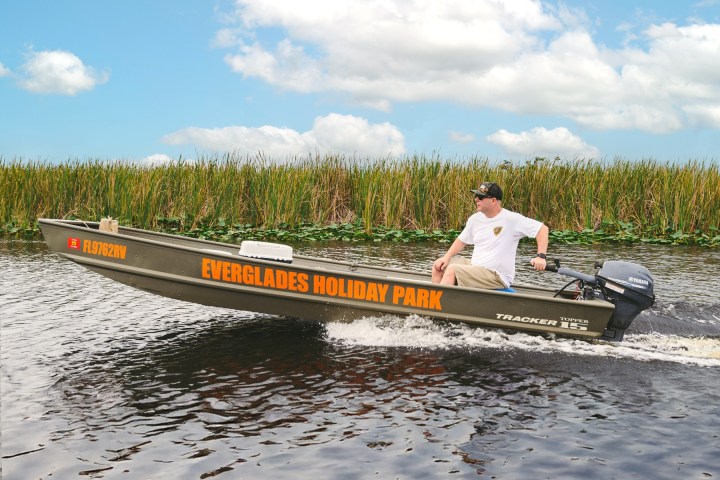 Person driving a motorboat labeled 'Everglades Holiday Park' on a waterway with reeds and a cloudy sky.
