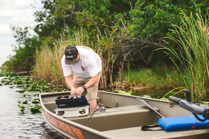 Person preparing fishing gear on a boat in a marshy area with tall grasses and trees.