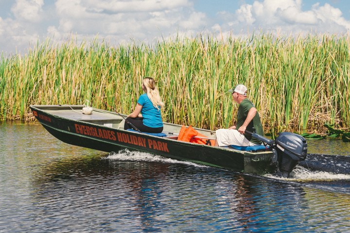 Two people on a motorboat navigating through tall grass in a waterway.