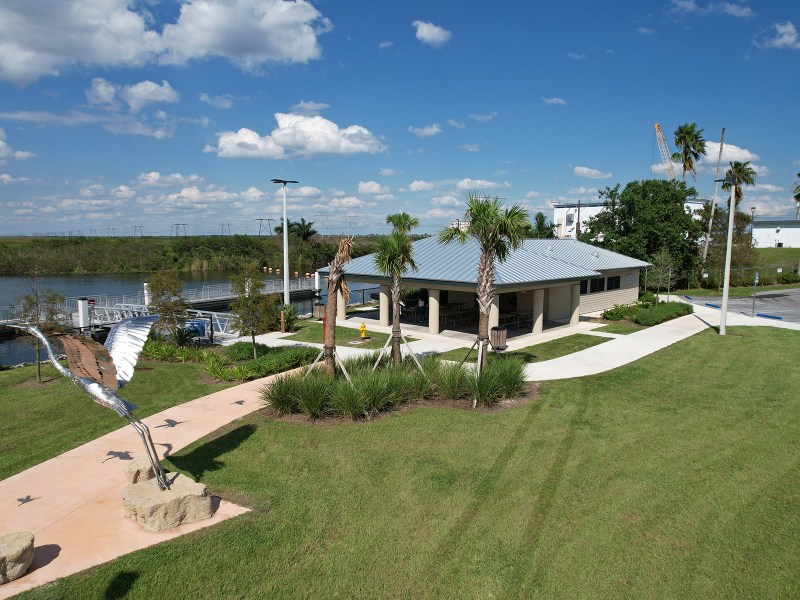 Small building, palm trees, river, and bird sculpture under a blue sky with clouds.