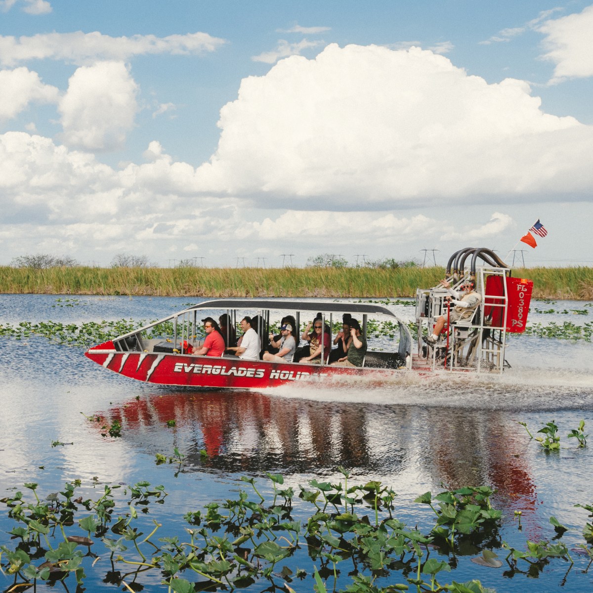 Airboat with passengers glides through marshy water with clouds overhead.