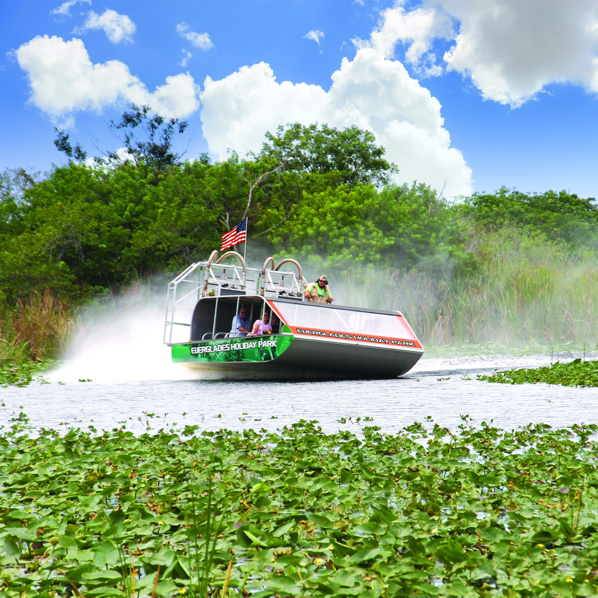 Airboat with American flag gliding over water in lush greenery under blue sky with clouds.