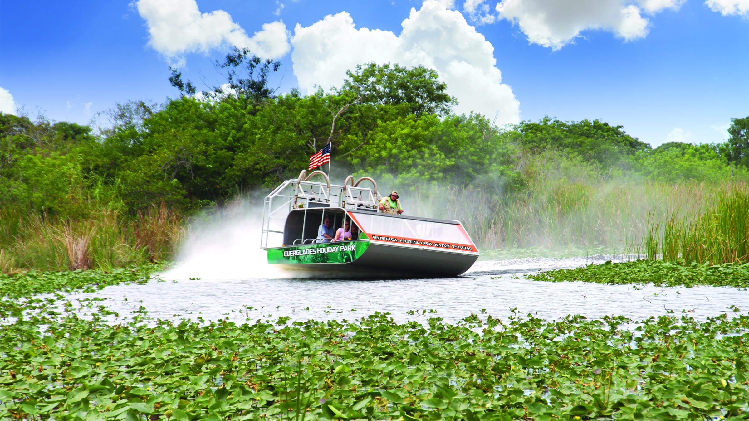Airboat with American flag gliding over water in lush greenery under blue sky with clouds.