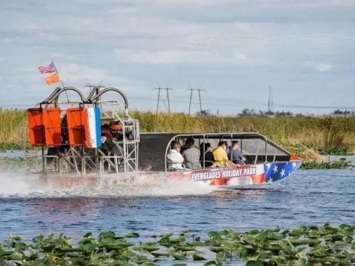 Airboat with passengers on a lake, American flag, lily pads, and grassy background.
