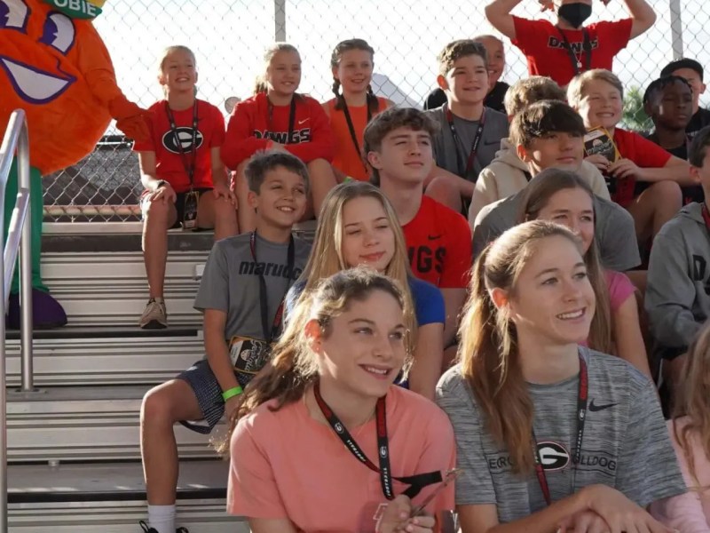 Group of smiling school kids on bleachers with an orange mascot beside them.
