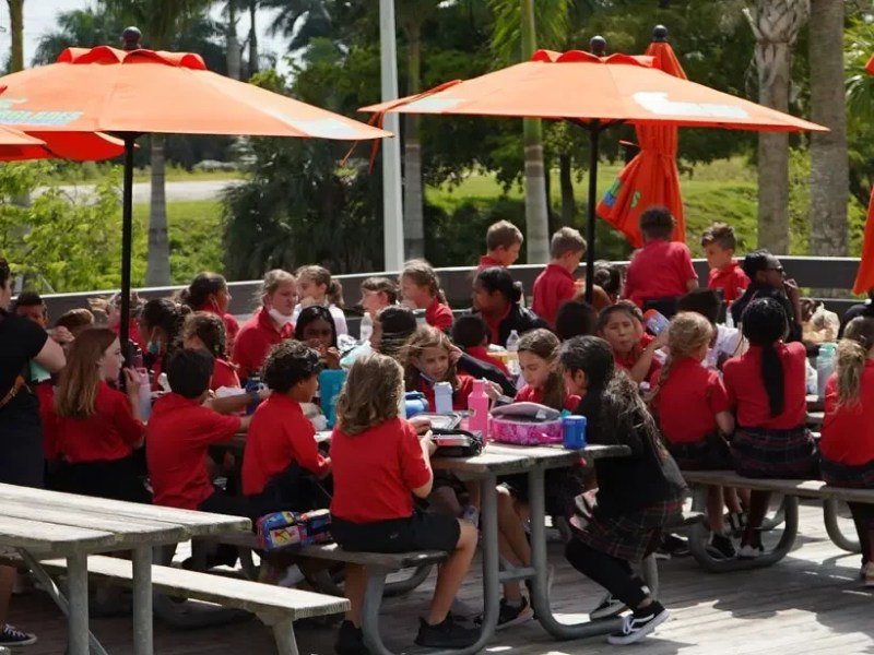 Children in red uniforms eating at outdoor tables with orange umbrellas.
