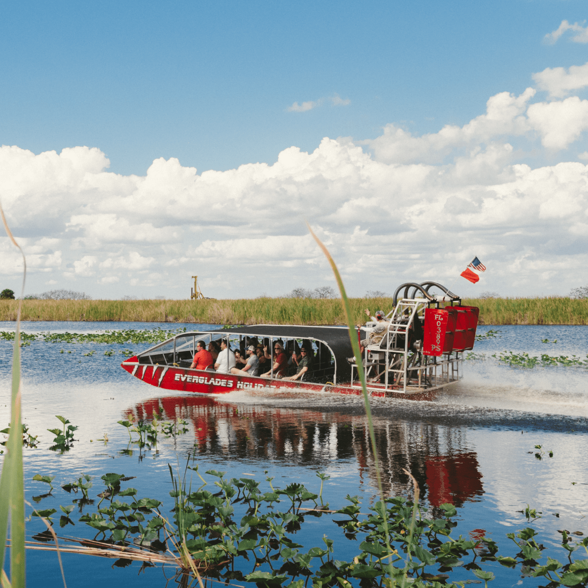 Airboat with tourists glides through water in a marshland under a blue sky with clouds.