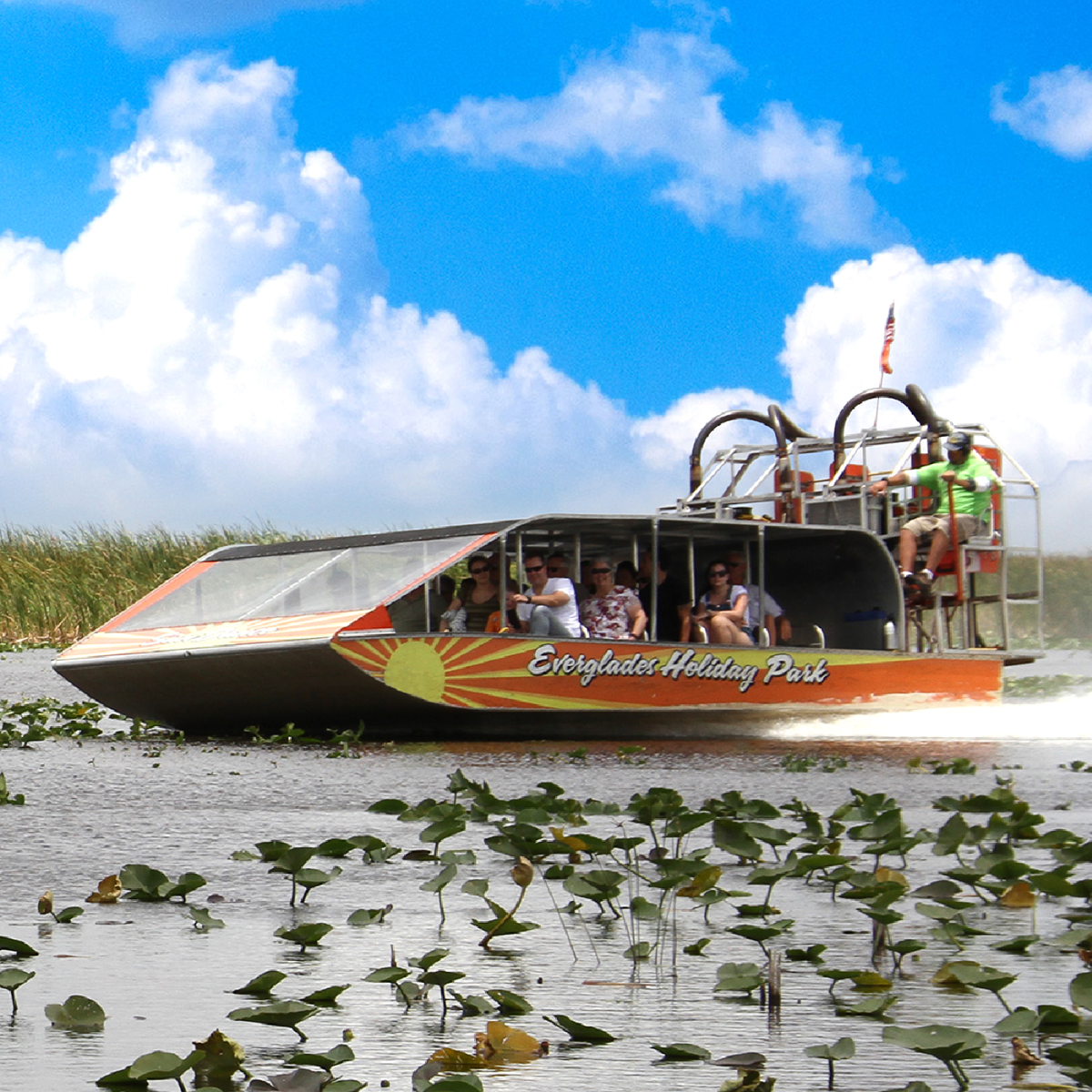 Airboat on water with passengers, surrounded by lily pads, under a blue sky with clouds.