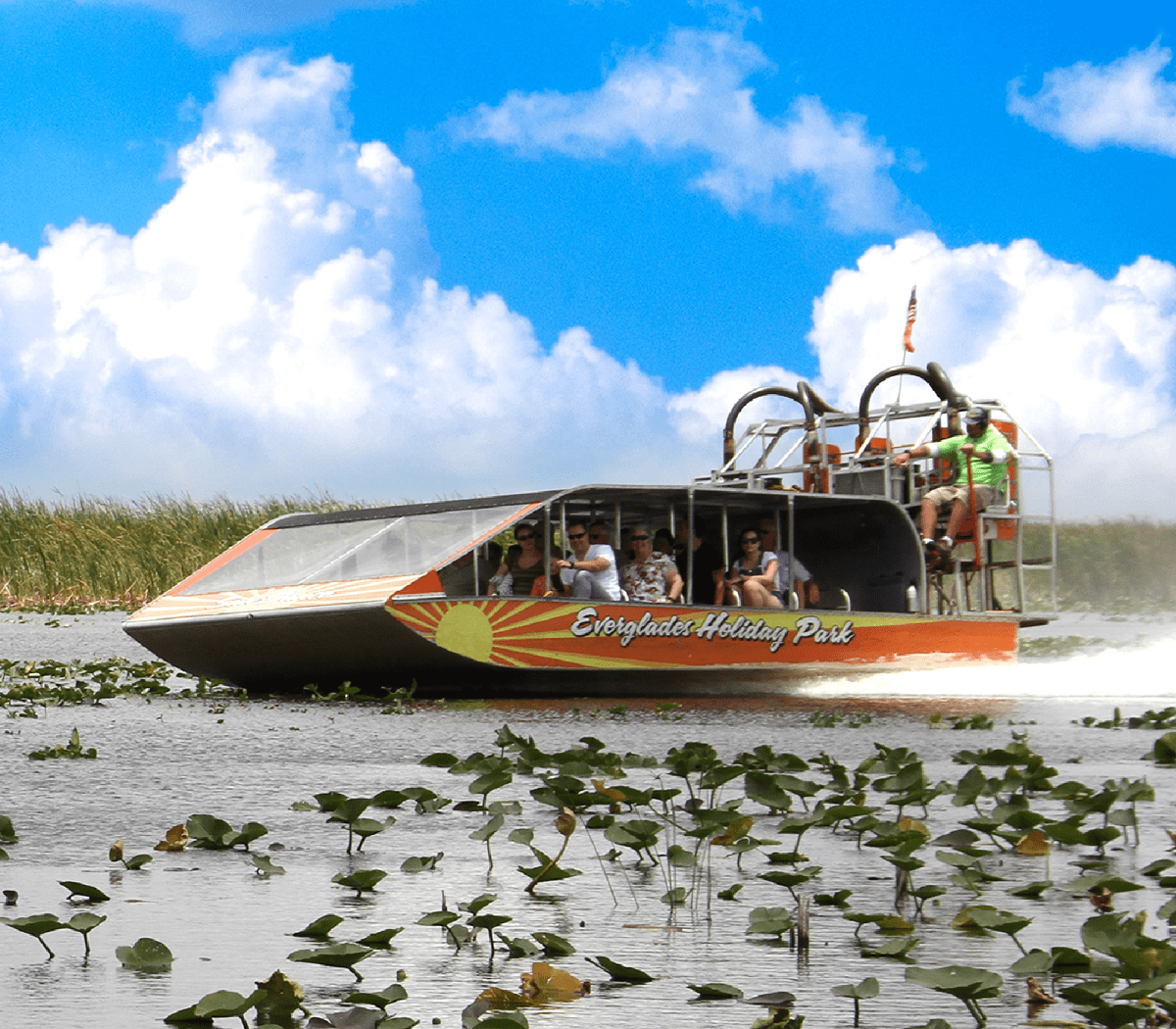 Airboat on water with passengers, surrounded by lily pads, under a blue sky with clouds.